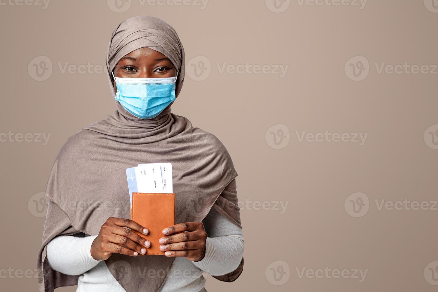 Traveling During Coronavirus Pandemic. Black muslim lady in protective medical mask holding passport and tickets, islamic woman in hijab posing over light studio background, ready for journey photo