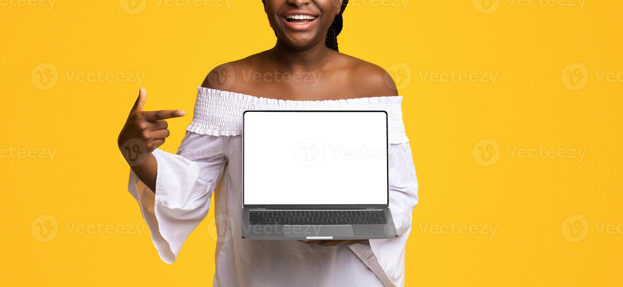 Check This Website. Cheerful Black Lady Pointing At Laptop Computer With Black Blank Screen, Standing On Yellow Background, Showing Free Place For Advertisement Or Design, Mockup With Copy Space photo