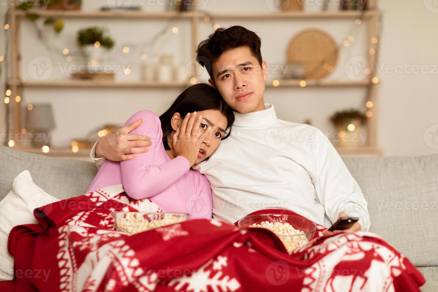Japanese Couple Watching Scary Movie On TV Spending Christmas Eve Sitting On Sofa In Front Of Television Set In Living Room At Home, Covered With Blanket. Family Watches Horror During Winter Holidays photo