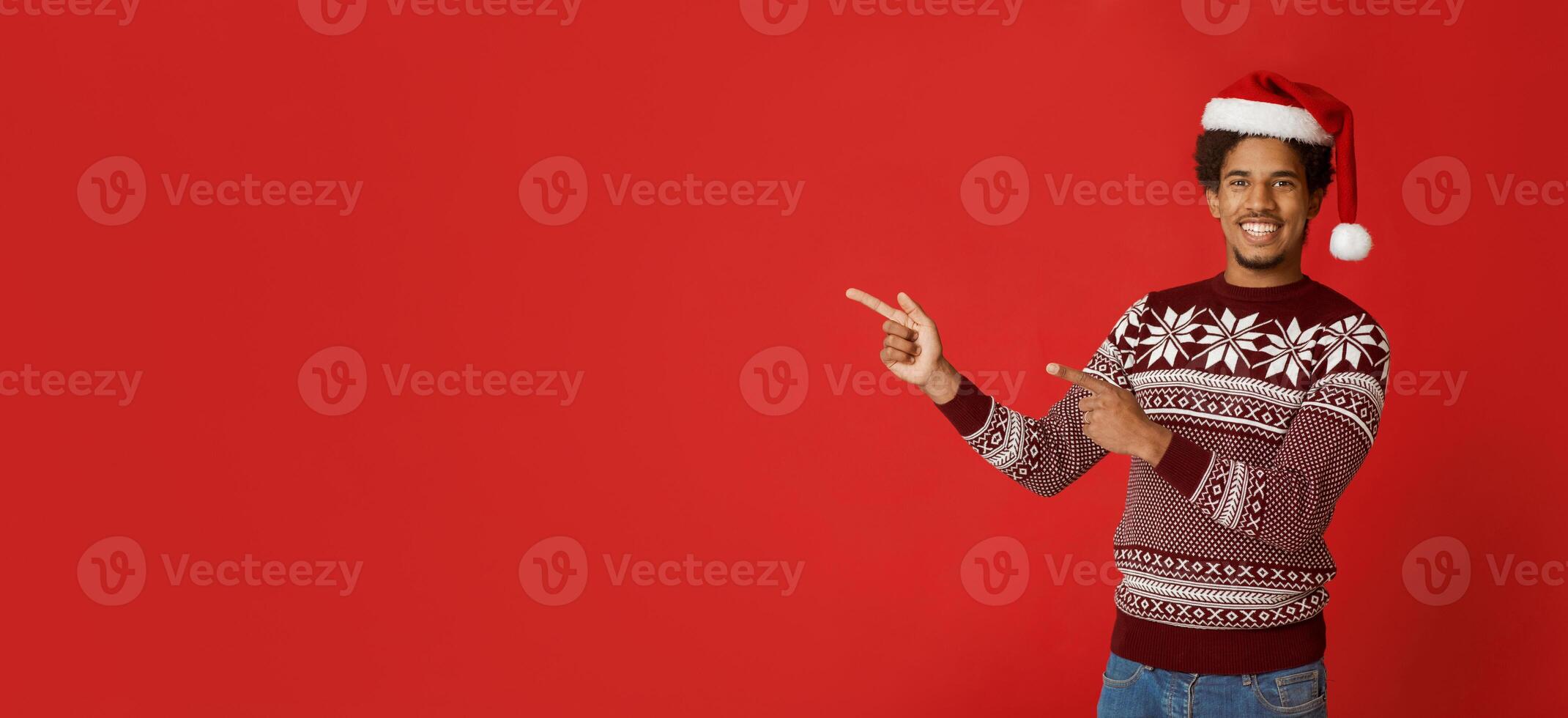 Best Christmas offer and seasonal sale or banner. Smiling african american millennial man in sweater and santa claus hat, points fingers to empty space, isolated on red background, studio shot photo