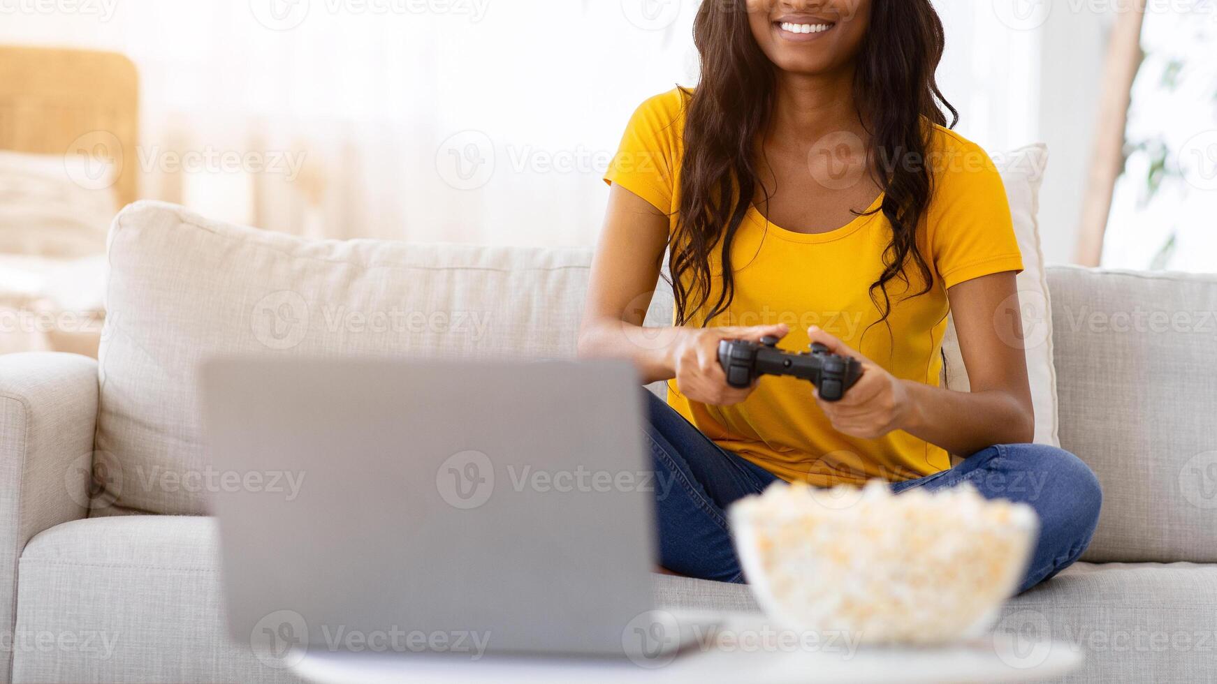 Pretty black woman enjoying videogame on playstation, resting at home. Joyful African American lady gamer with joystick and popcorn sitting on couch, playing computer arcade photo