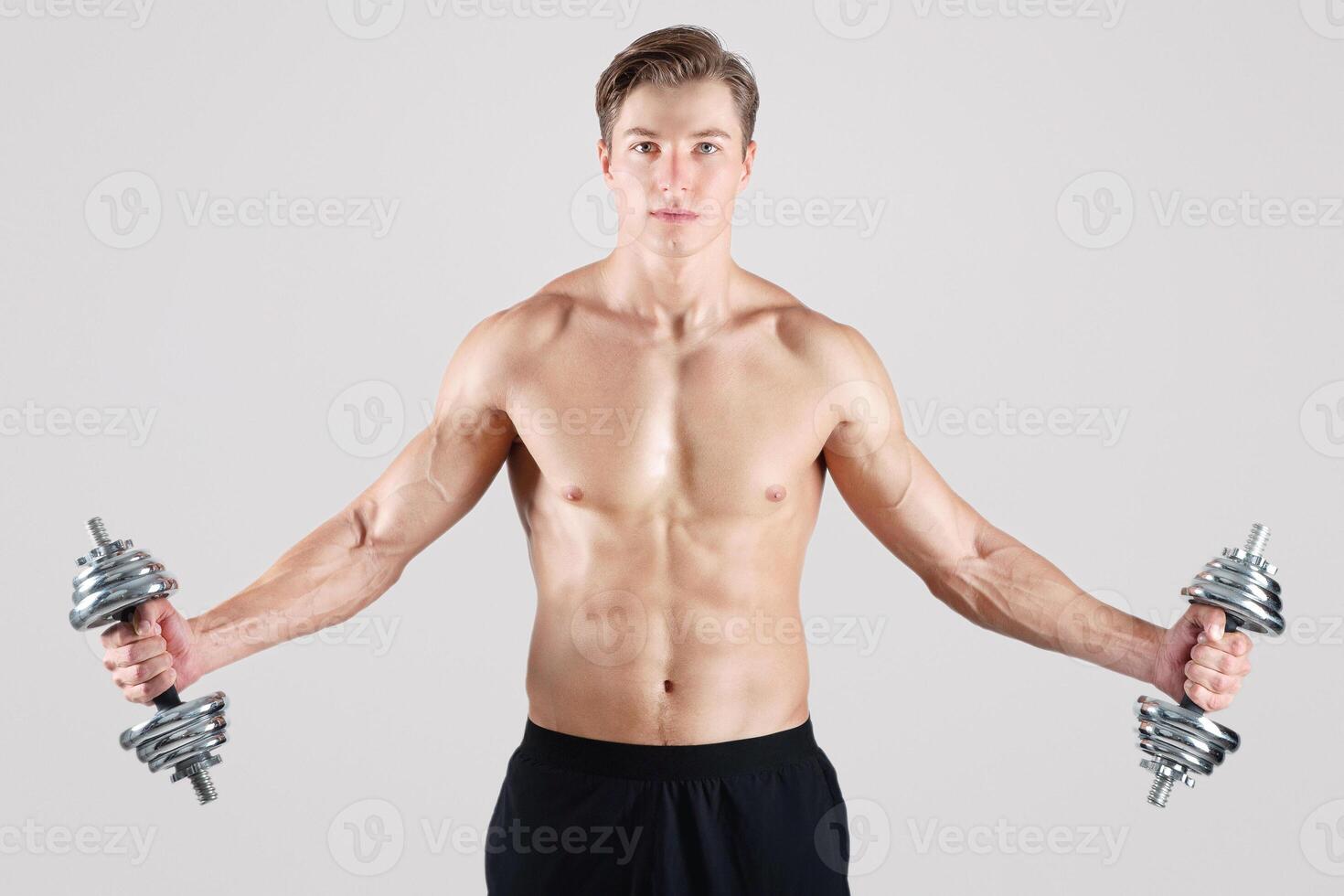 Strength training concept. Fit young guy with naked torso and six pack abs lifting dumbbells over light studio background. Handsome muscular bodybuilder working out with weights photo