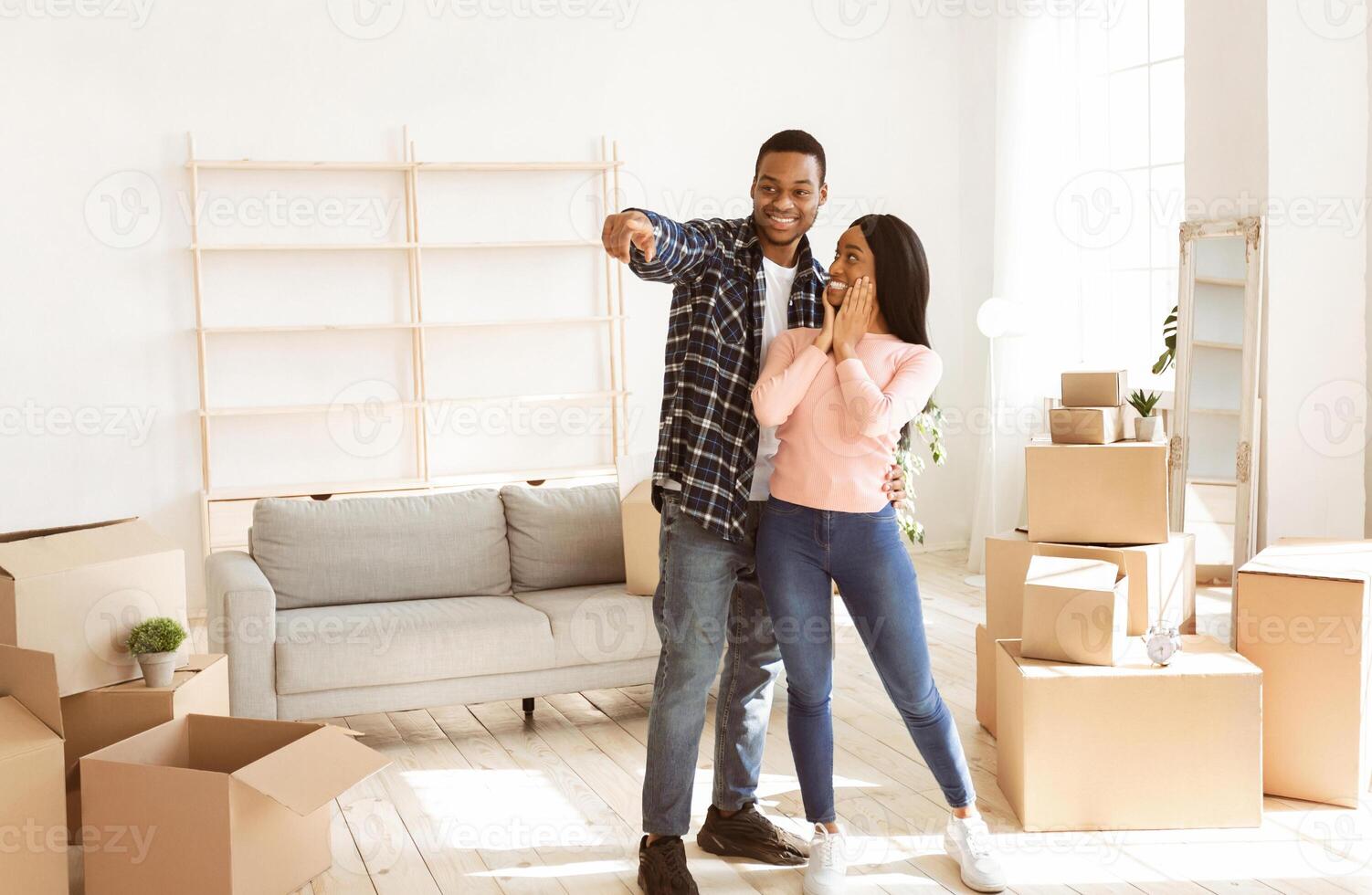 Black millennial man showing something to his excited girlfriend in their new house, copy space. Smiling African American guy pointing at place to put furniture. Moving day concept photo