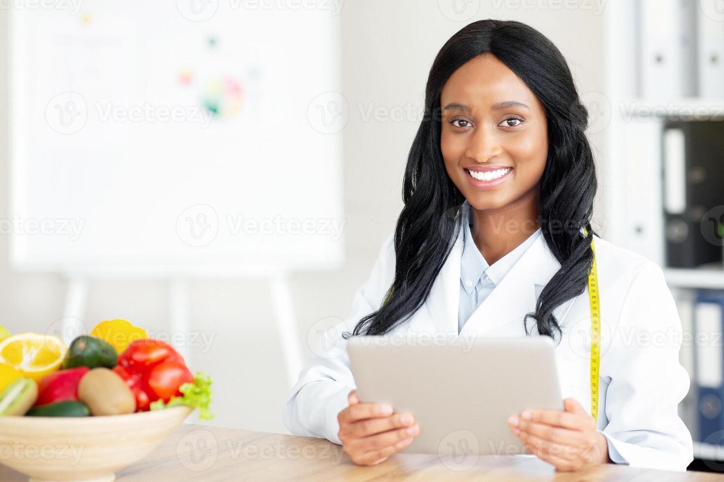 Young black nutritionist with touch pad creating plant based meal plan for client at clinic, blank space. Joyful female dietitian with tablet consulting patient about healthy diet online at office photo