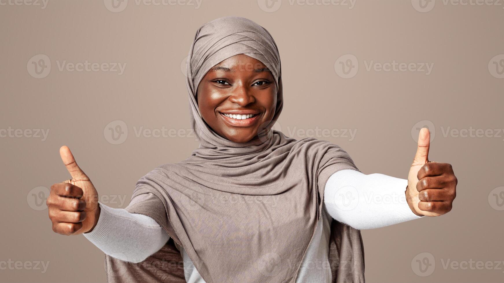 Like. Joyful black muslim lady in headscarf showing thumbs up at camera, smiling african woman in hijab recommending great offer, gesturing sign of approval, standing over light studio background photo