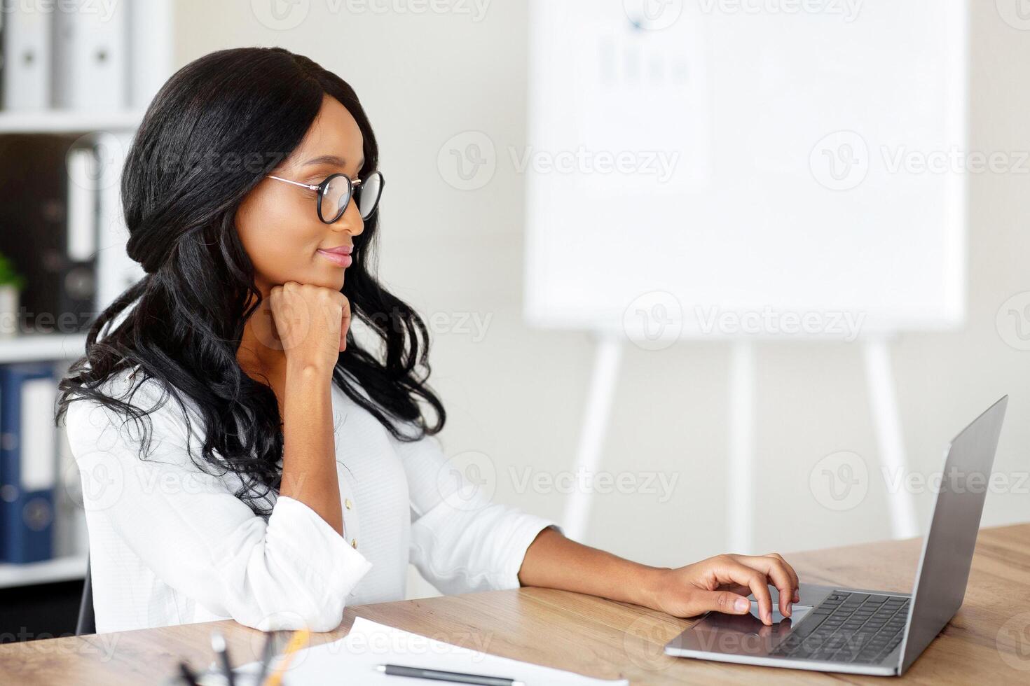 Positive black business lady using laptop pc, working with business correspondence in her office, copy space. Attractive African American employee making online business call or presentation photo
