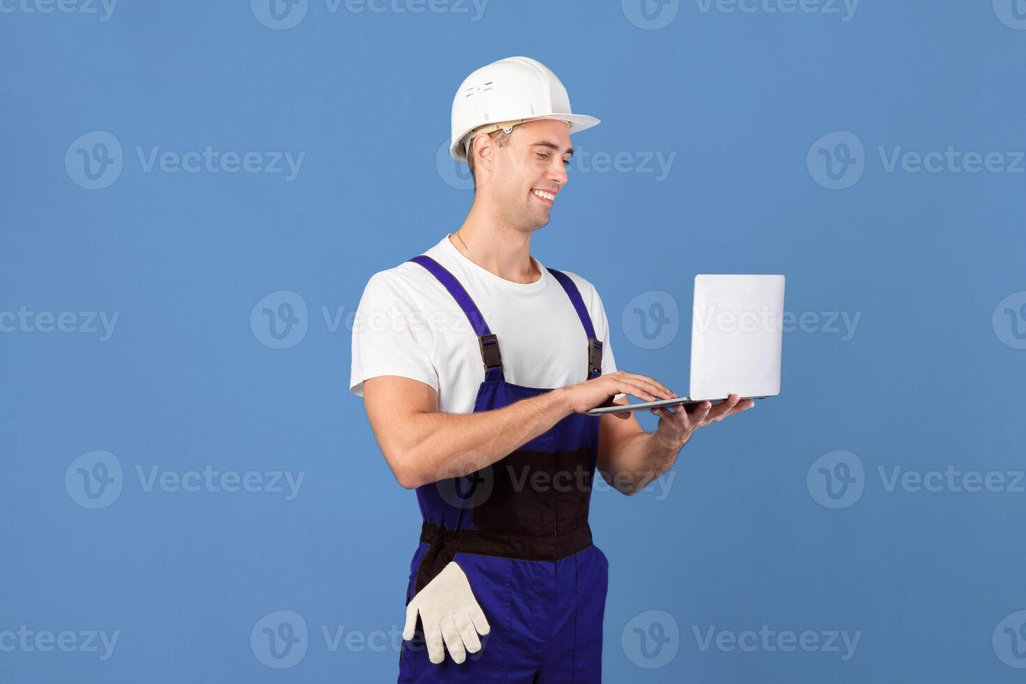 Handsome Young Foreman Guy With Laptop, Ordering Construction Materials Online. Male Worker In Hardhat Buying Repair Tools In Internet, Shopping On New Website, Standing On Blue Background, Copy Space photo