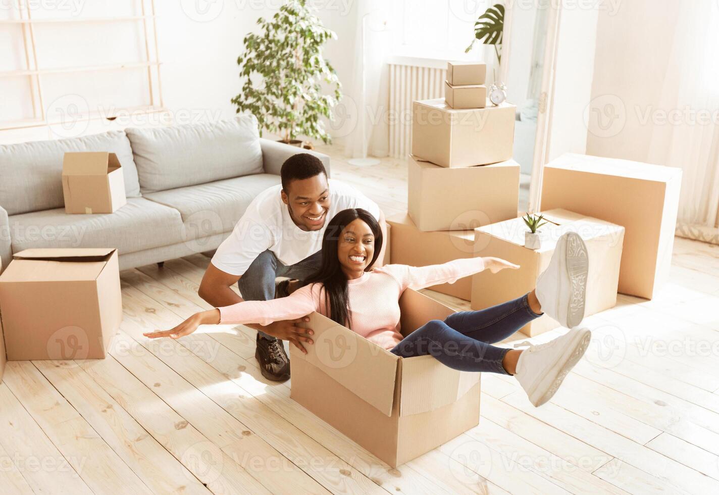 Beautiful black lady and her young husband playing together with cardboard box on moving day. Positive African American family entertaining themselves in their new house during relocation photo