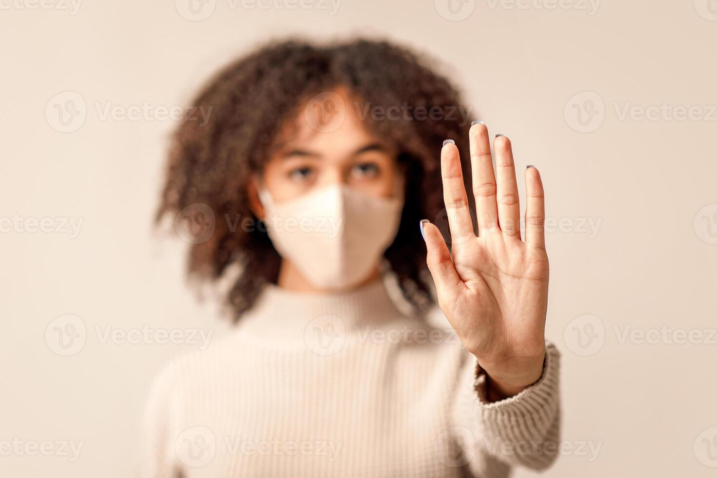 Social distance and fight against coronavirus during pandemic. Focus on hand of serious african american woman in protective mask and sweater, isolated on light background, studio shot, free space photo
