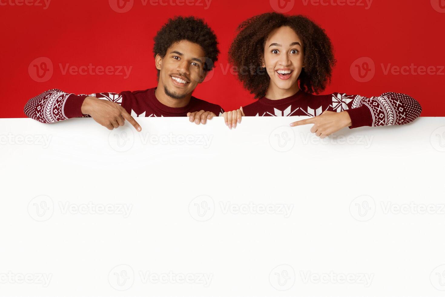 Happy New Year and merry Christmas. Laughing young african american couple in love in identical sweaters over white blank billboard and point fingers on it, isolated on red background, studio shot photo