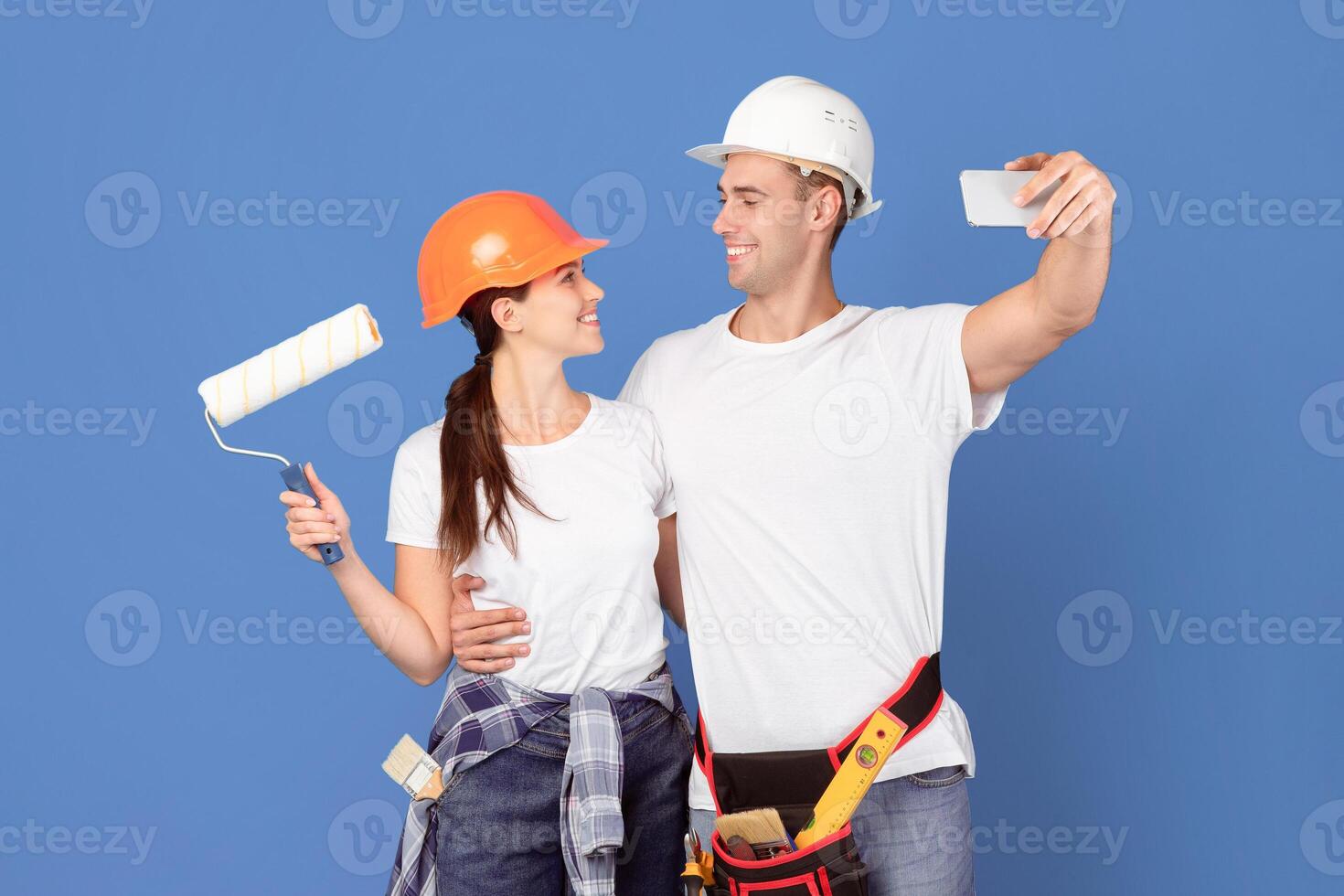 Cheerful Repair Workers Couple In Protective Hard Hats Taking Selfie On Smartphone, Posing Over Blue Background, Ready For House Renovation. Man Wearing Tool Belt And Woman Holding Paint Roller photo