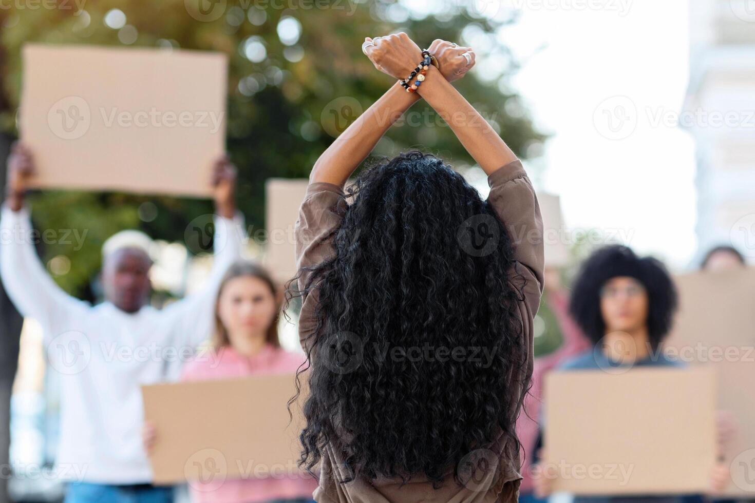 Unrecognizable long-haired brunette woman leading group of protestors holding blank placards, back view of lady standing in front of multiracial demonstrators, raising fists up and crossing hands photo