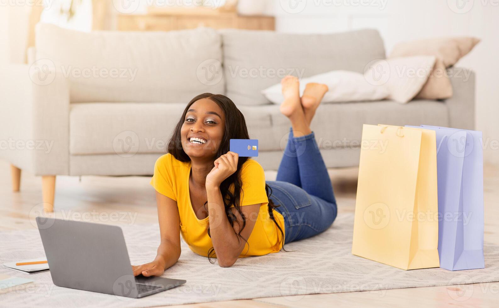 Happy African American woman with laptop computer and credit card doing online shopping at home. Smiling black lady making purchase via internet while lying on floor indoors. Panorama photo