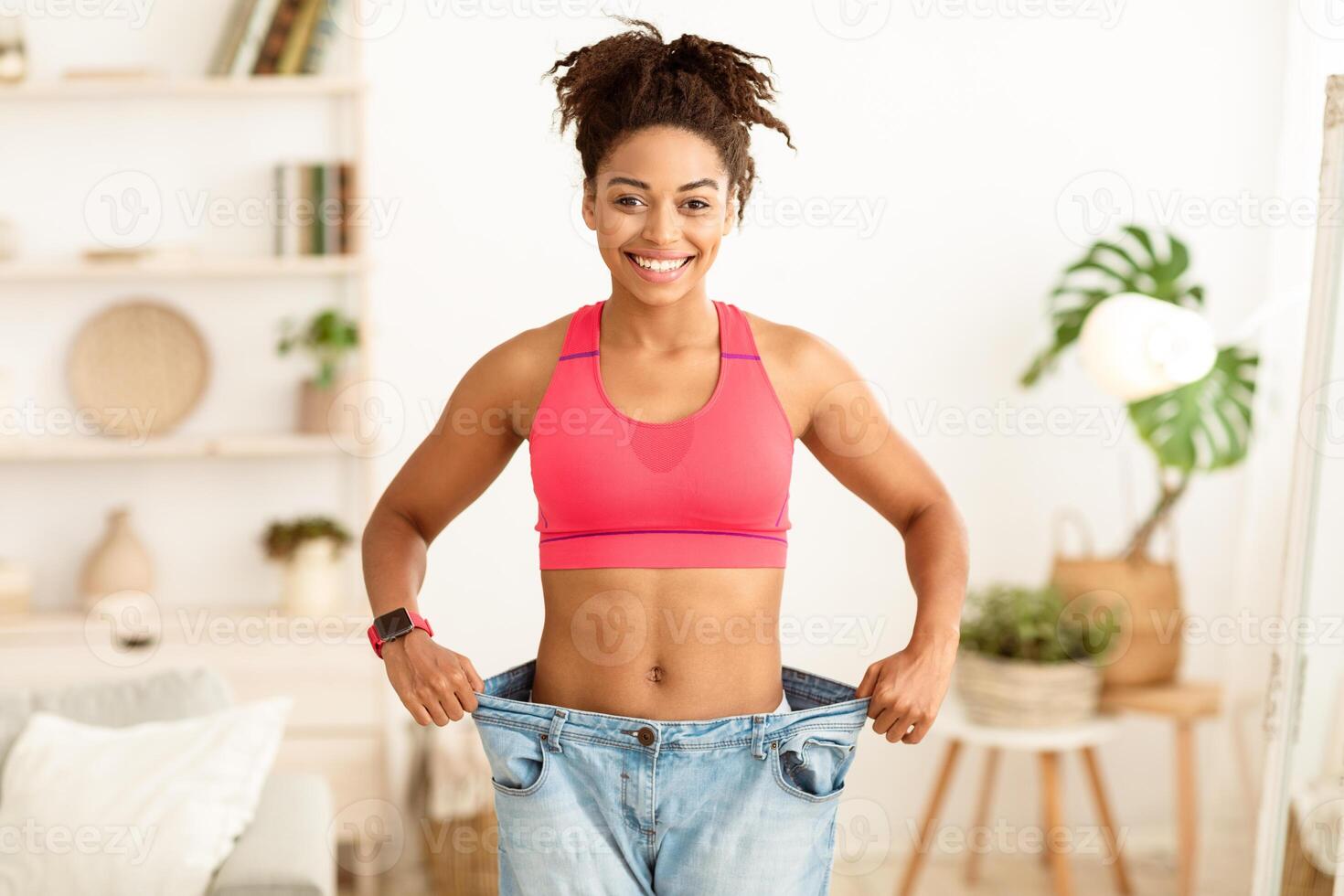 Great Weight Loss. Joyful Slim Black Girl Wearing Old Big Jeans Showing Result Of Losing Excess Kilograms Standing Indoors, Smiling To Camera. Dieting And Slimming Motivation Concept photo