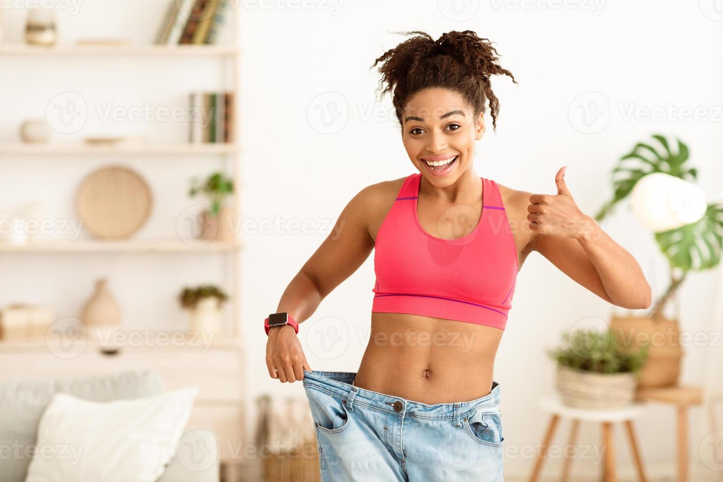 Cheerful African American Woman Showing Result Of Weight Loss Gesturing Thumbs Up Approving Diet, Wearing Old Oversize Jeans After Slimming Standing At Home, Smiling To Camera. Empty Space For Text photo