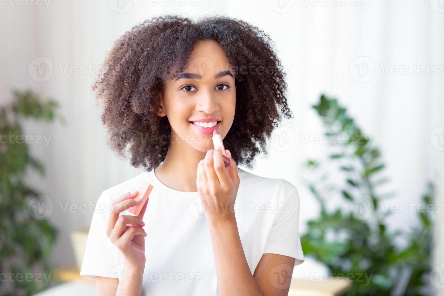Moisturizing lips and prevention of peeling cracking of delicate skin. Millennial african american female makes nude makeup, use professional decorative cosmetic, lipstick on lips in living room photo
