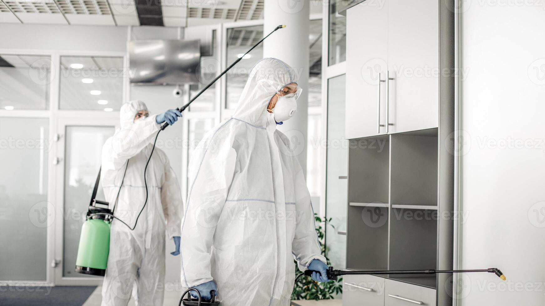 Professional disinfection of room and furniture during coronavirus epidemic. Men in hazmat suits, protective masks, goggles and with sprayer, cleans office with chemical, free space photo