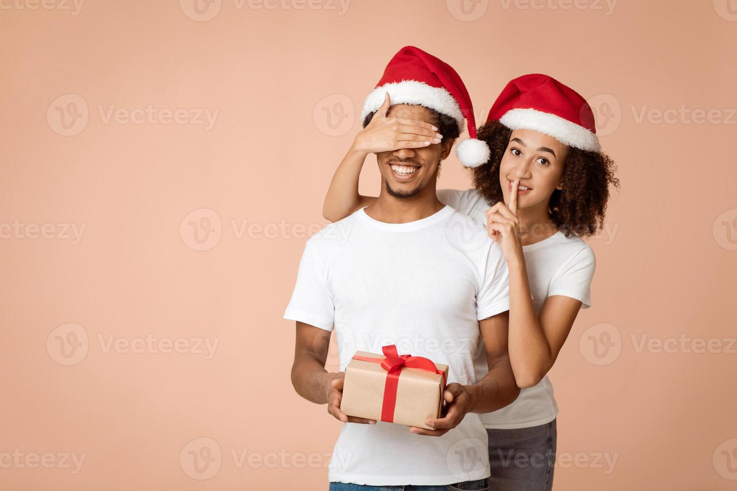 Romantic Xmas and New Year together. Cheerful african american lady in Santa hat shows sign of silence with hand and closes eyes to husband with gift box on light background, empty space, studio shot photo