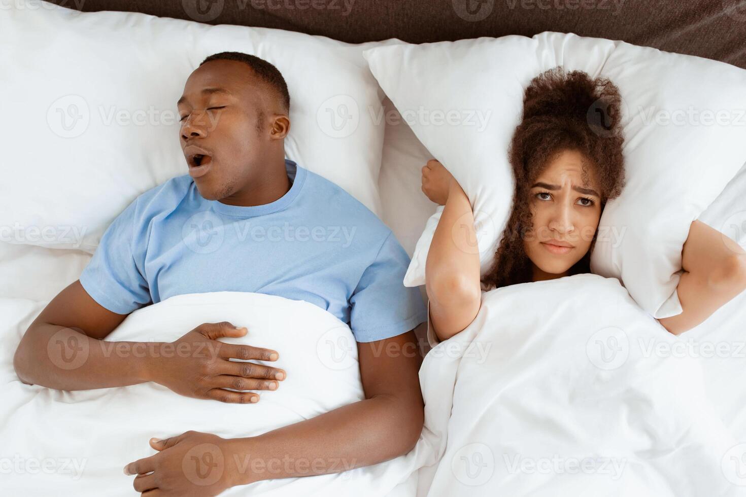 Snoring man sleeping with apnea and sleepless woman, angry wife unable to get to sleep. Upset young african american lady covers ears with pillow so as not to hear noise from snoring guy in bedroom photo