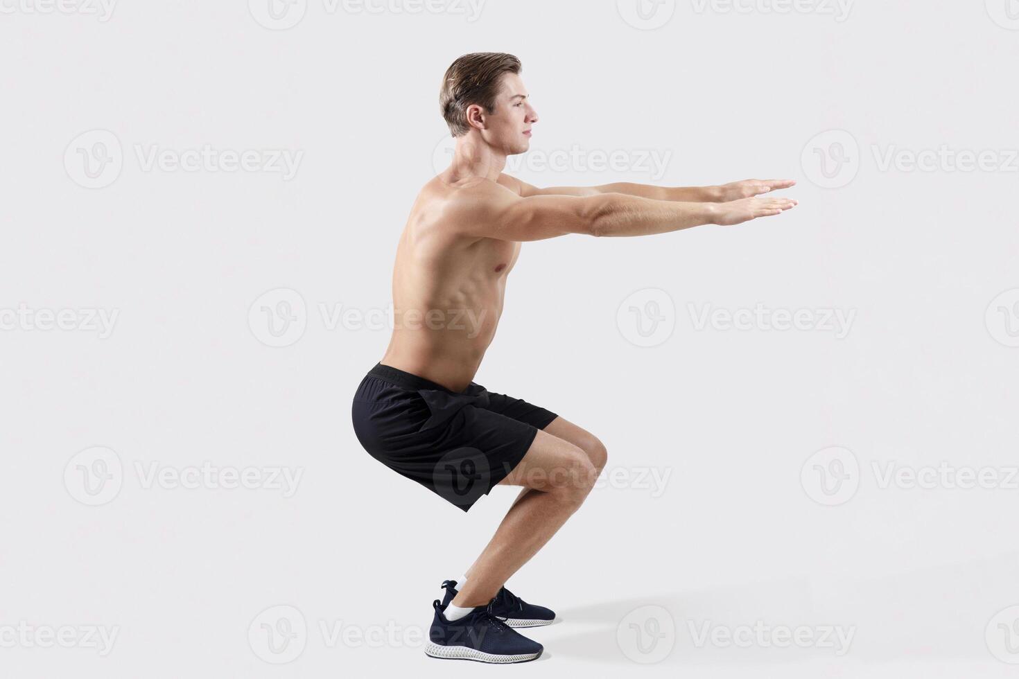 Side view of shirtless athletic man with bare torso doing aerobic exercises on light studio background. Sexy young guy with muscular body making squats, full length portrait. Sports concept photo