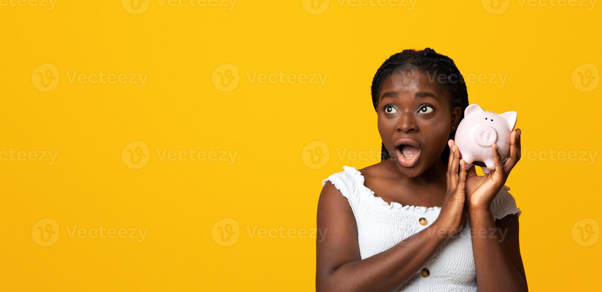 Shocked african american woman listening sound of coins in piggy bank, checking saved money, planning budget for future, emotional black lady standing over yellow studio background with copy space photo