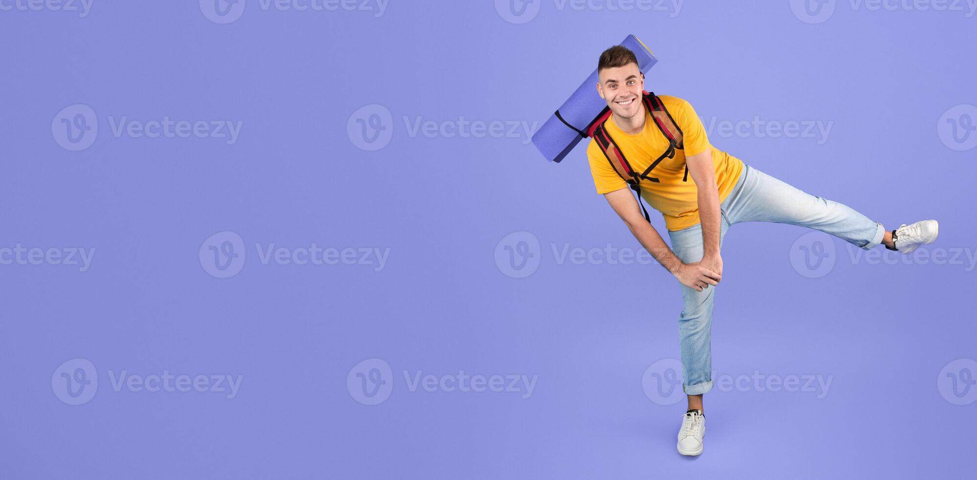 Full length portrait of happy young man with camping equipment dancing over blue studio background. Millennial male traveler excited to go hiking, trekking or mountaineering. Tourism concept photo