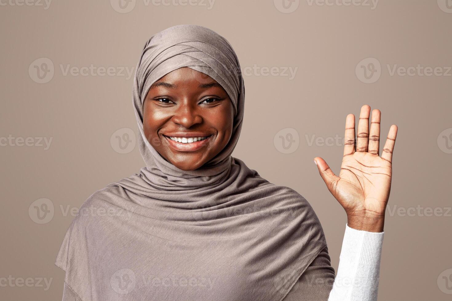 Hello Gesture. Friendly Black Muslim Lady In Headscarf Waving Hand At Camera, African Islamic Lady Gesturing Hi With Open Palm, Greeting Someone, Posing Over Light Background In Studio photo