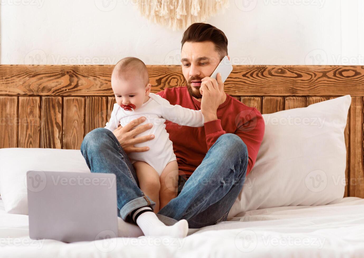 Father Talking On Phone Caring For Little Baby And Working On Laptop Distantly Sitting In Bedroom At Home. Daddy Works And Spending Time With Toddler Child During Paternity Leave photo