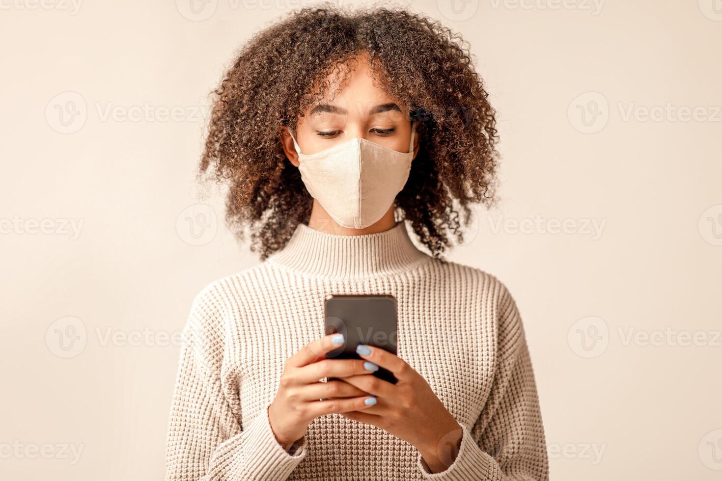 Remote communication online during spread of virus and social distance. Young african americans lady in protective mask and sweater surfing on smartphone isolated on light background, studio shot photo