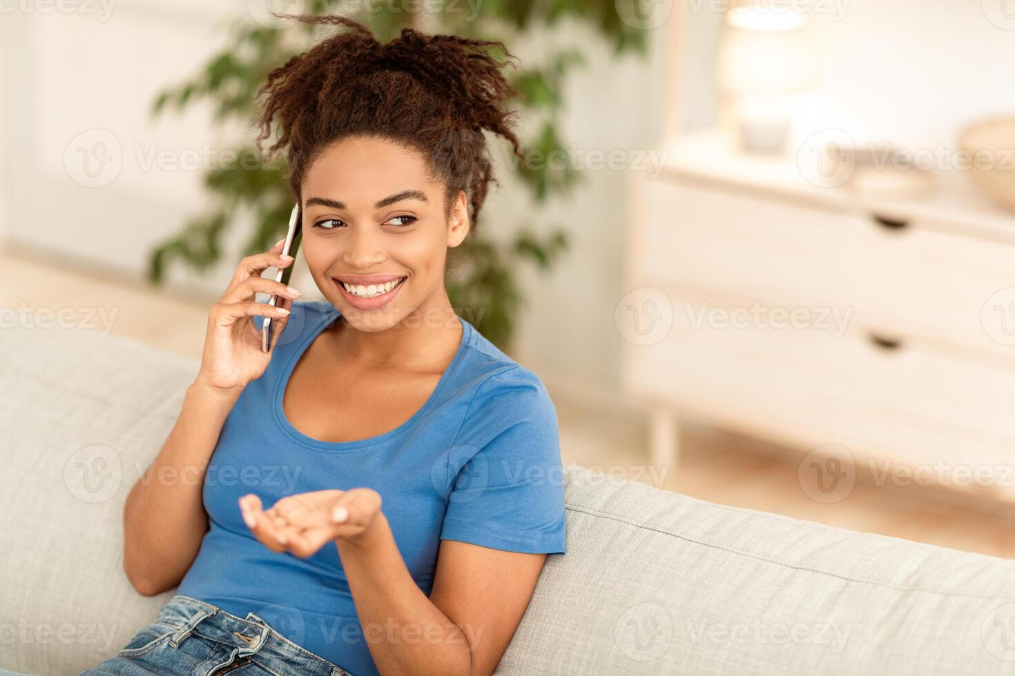 Cheerful Black Young Woman Talking On Cellphone Sitting On Sofa At Home, Chatting And Discussing News With Friend. Phone Call, Distant Communication Concept. Empty Space For Text photo