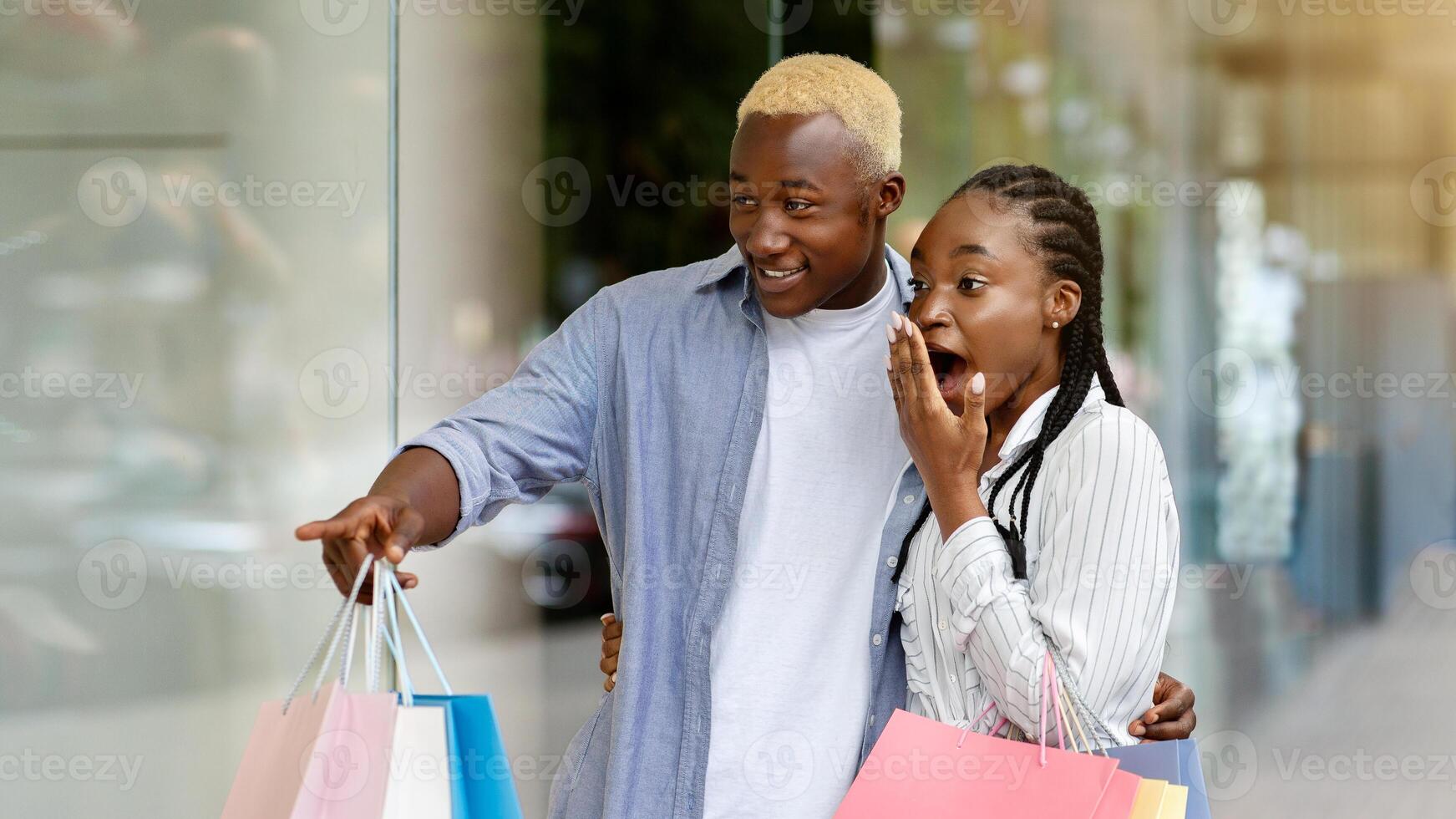 I want it now. Shocked african american young man points at shop window, to girl with colored bags, free space photo