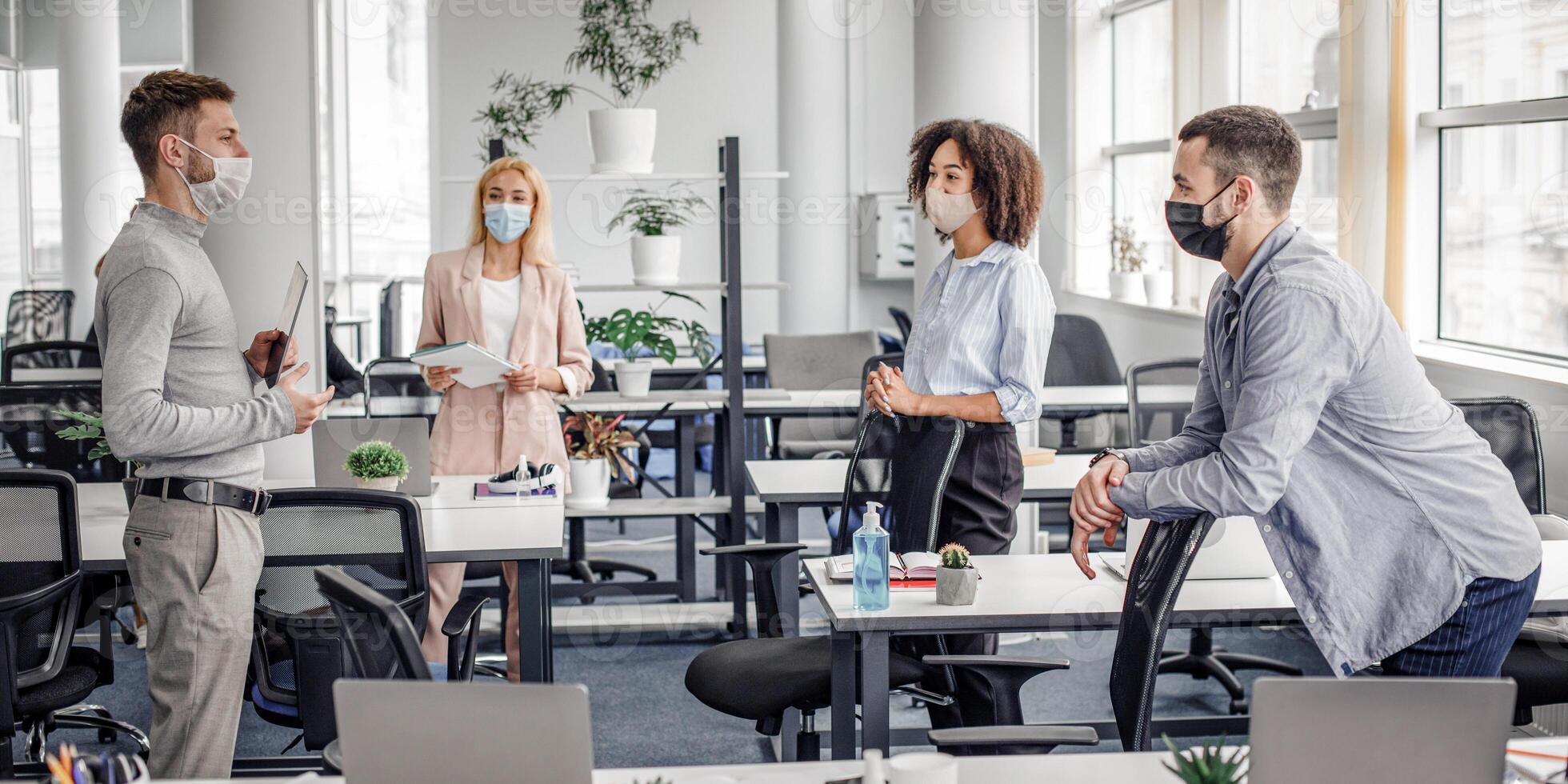 Modern team meeting, group work and social distancing. Manager with tablet speaks with workers in protective masks in interior of modern office with gadgets during coronavirus epidemic, free space photo