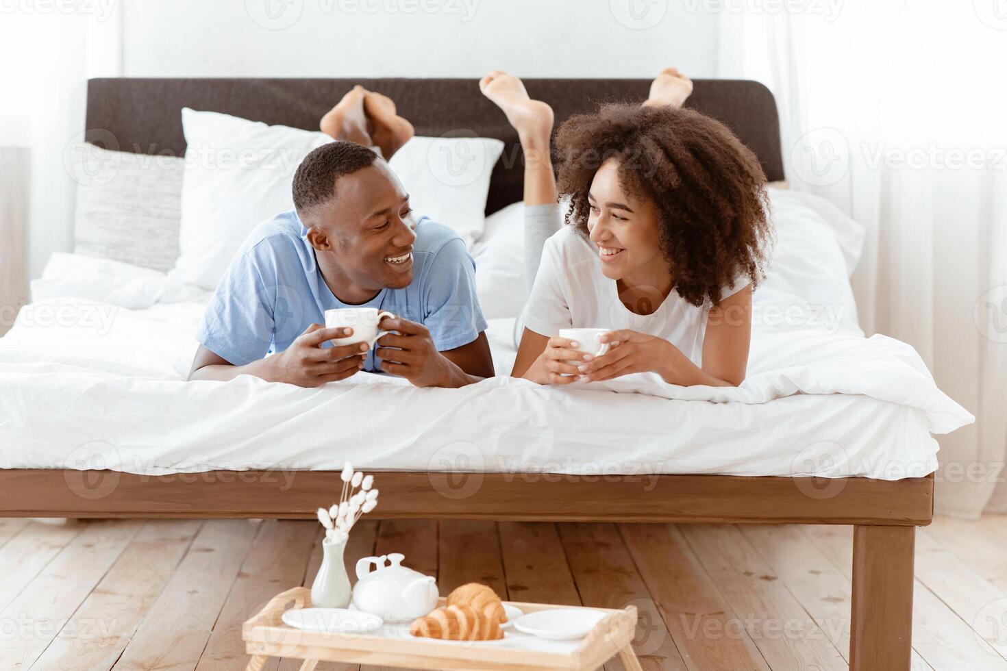 Free time in morning or weekend, breakfast in bed together and self-isolation at home. Cheerful millennial african american male and female lie, talk, drink hot tea, with tray of croissants in bedroom photo