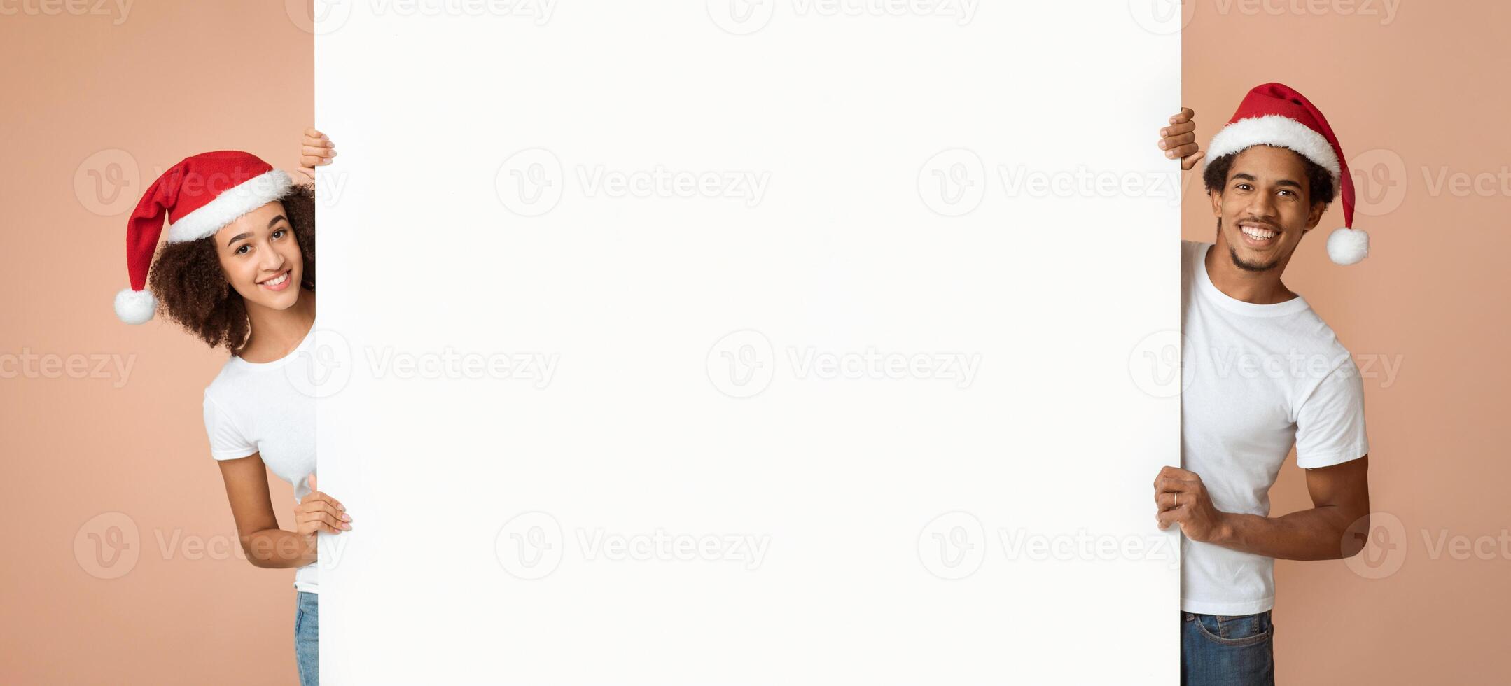Christmas and New Year with family. Funny millennial african american guy and lady in santa claus hats holds large empty billboard for word, look at camera, isolated on light background, studio shot photo
