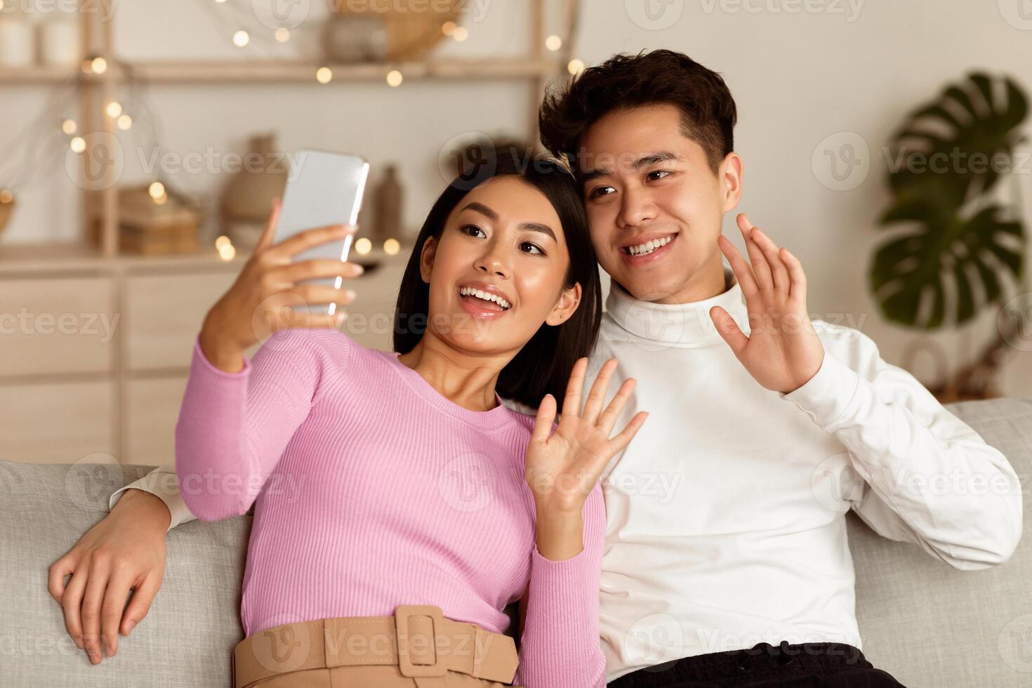 Asian Family Couple Making Call Using Smartphone Waving Hello Sitting On Couch At Home. Married Spouses Calling Distant Parents, Remote Communication With Relatives And Friends Concept photo