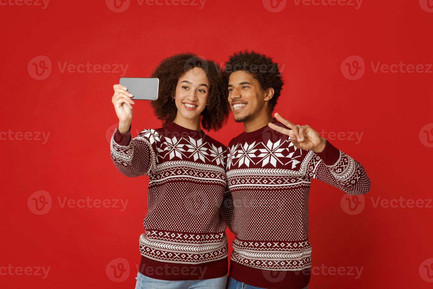 Couple of lovers in red Christmas clothes making selfie photo. Young african american man and woman looking at camera of smartphone, male makes peace sign with his hand, isolated on red background photo