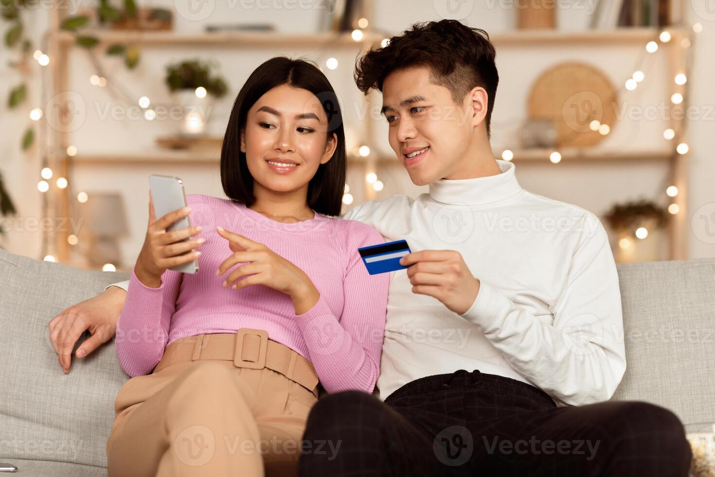 Mobile Banking App. Asian Couple Shopping Online With Smartphone And Credit Card Choosing And Buying Things In Internet Shop On Phone Sitting On Sofa In Cozy Living Room At Home. Selective Focus photo