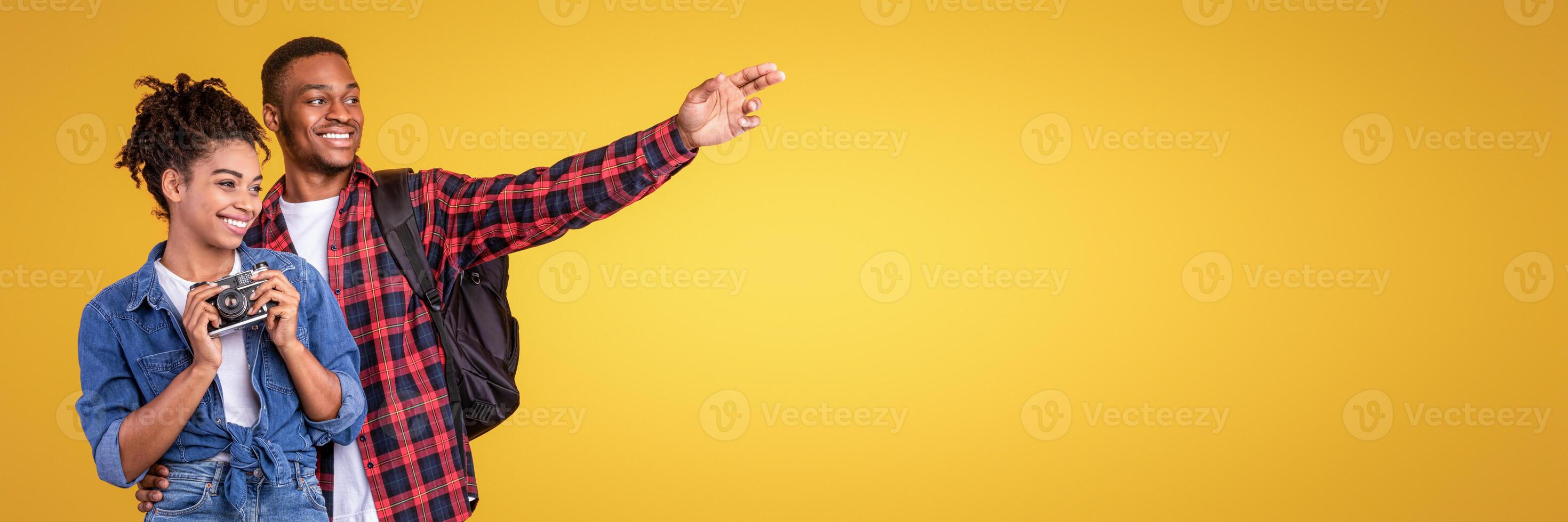Travel Photography. Smiling Black Tourists Couple Holding Photo Camera Looking Aside And Pointing Fingers Standing On Yellow Studio Background. Vacation And Tourism Concept. Panorama With Copy Space