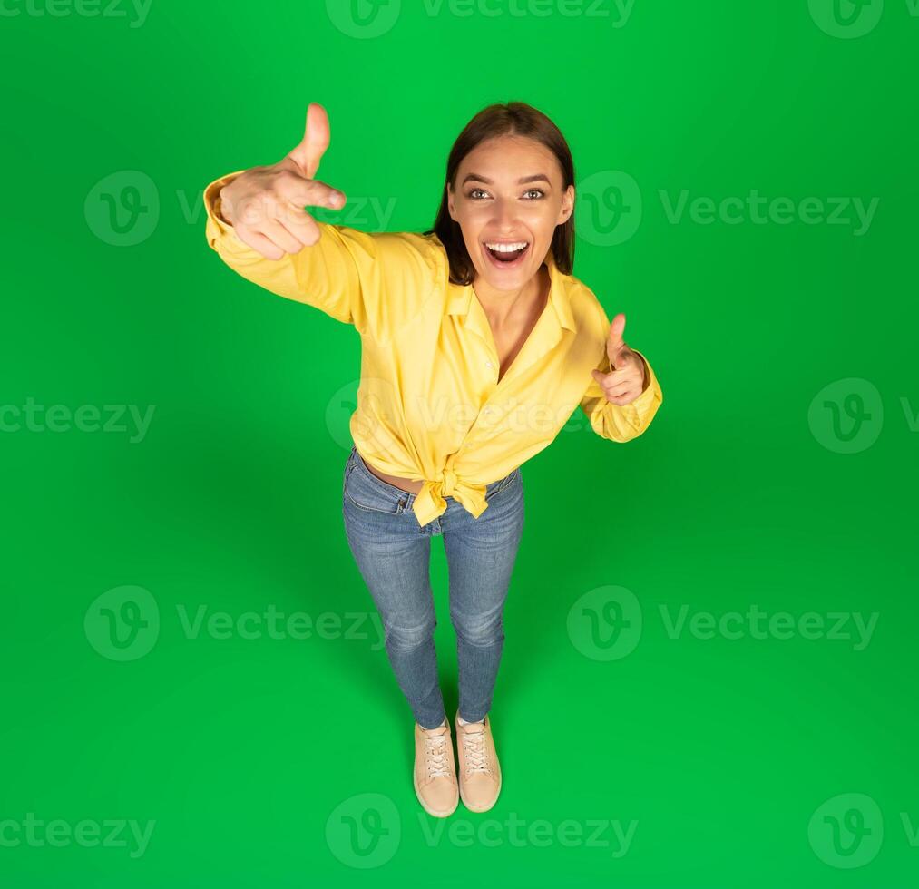 Hey, You. Top view portrait of funny excited young woman pointing fingers at camera posing standing in studio on greeb background. I choose you concept, advertisement banner with cheerful casual lady photo
