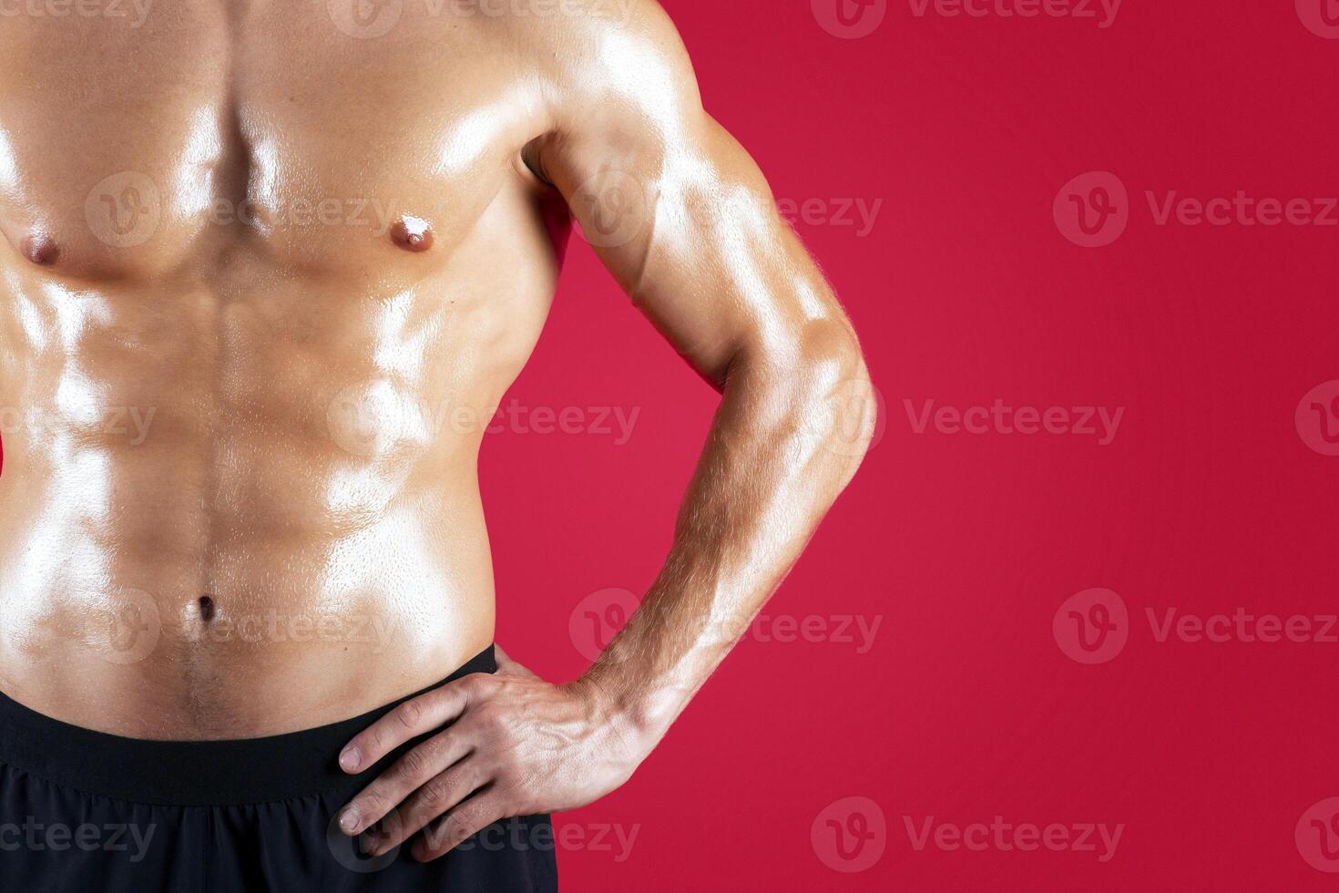 Closeup of sexy young guy with naked torso posing with hands on hips on red studio background, copy space. Cropped view of handsome fit man with six pack abs. Healthy living concept photo