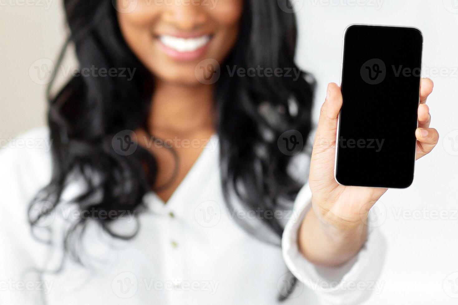 Cheerful black businesswoman holding smartphone with blank screen at office, mockup for mobile app or website design. Successful female manager with cellphone, space for your ad. Selective focus photo