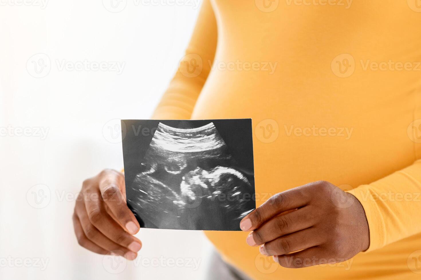 Unrecognizable pregnant african american woman holding ultrasound image of her unborn child, standing by window at home, copy space. Cropped of black lady with big tummy showing sonogram photo