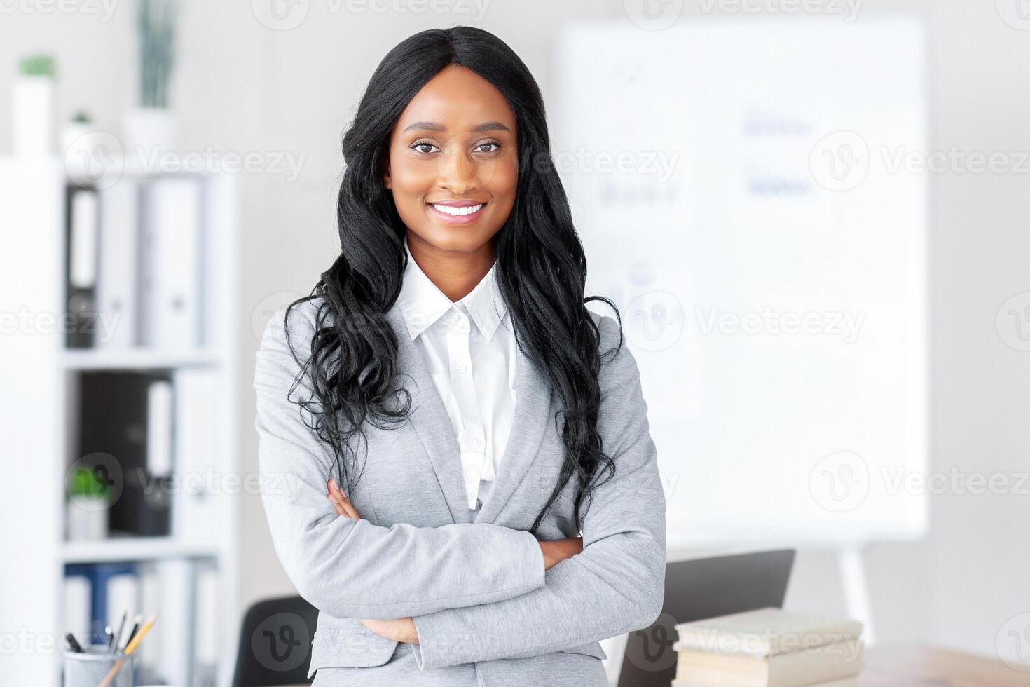 Portrait of friendly black female manager in formal suit standing with crossed arms at contemporary office. Successful African American busineswoman posing and smiling at camera photo