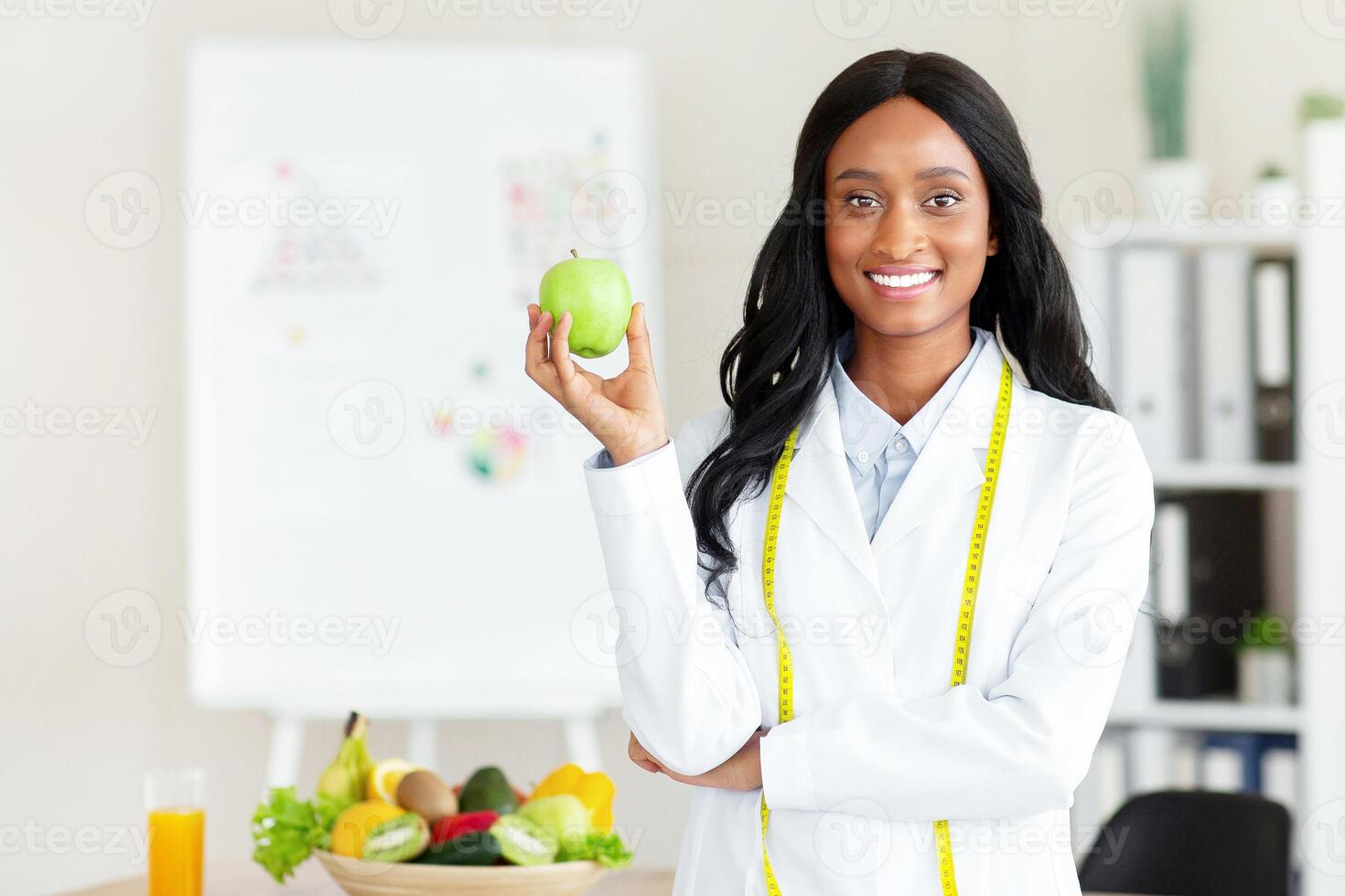 Smiling black female nutrition adviser with apple and tape measure looking at camera at clinic. Weight loss specialist promoting healthy eating habits at medical office photo