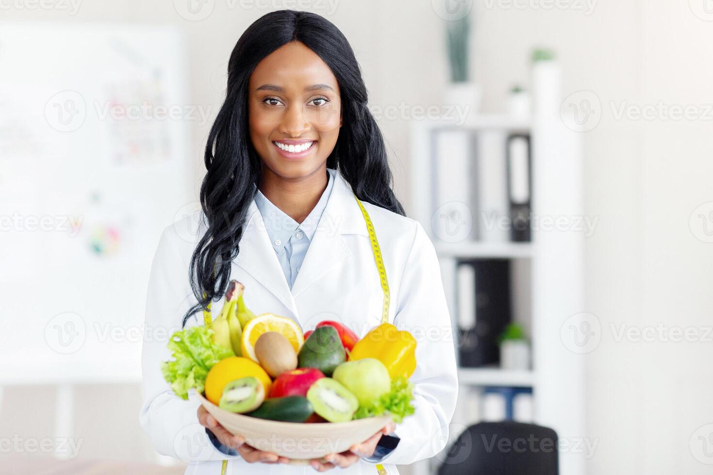 Portrait of happy black dietitian in lab coat holding bowl of fresh fruits and vegetables, smiling at camera at clinic. Weight loss consultant recommending healthy plant based diet photo