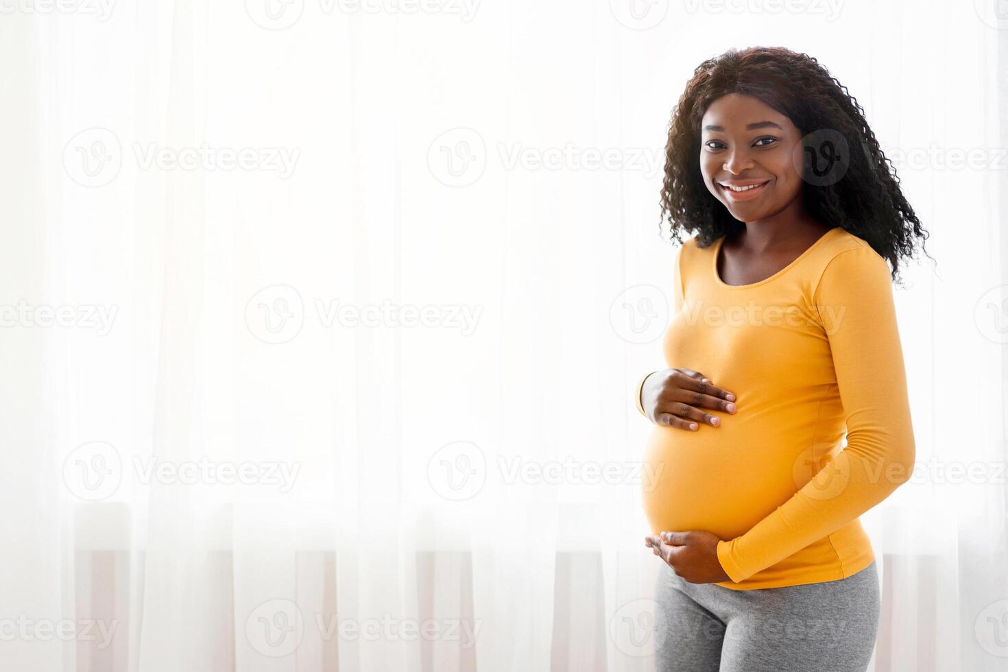 Beautiful pregnant african american woman hugging her big belly, enjoying her pregnancy, free space. Side view of young black expecting lady standing next to window at home, touching her tummy photo