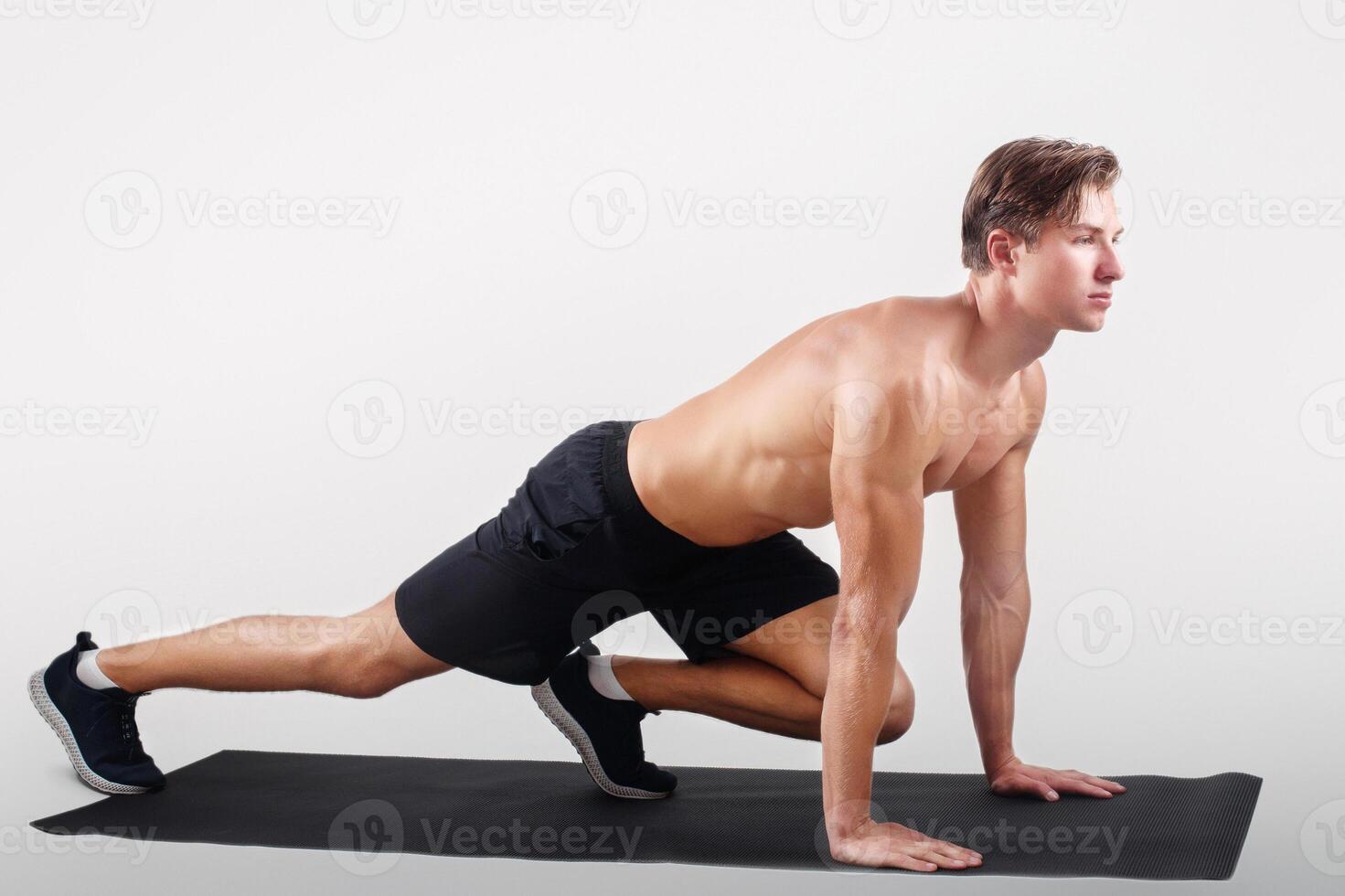 Healthy lifestyle and sports concept. Strong young man with naked chest exercising on yoga mat over light studio background. Motivated millennial sportsman doing pilates, full length photo