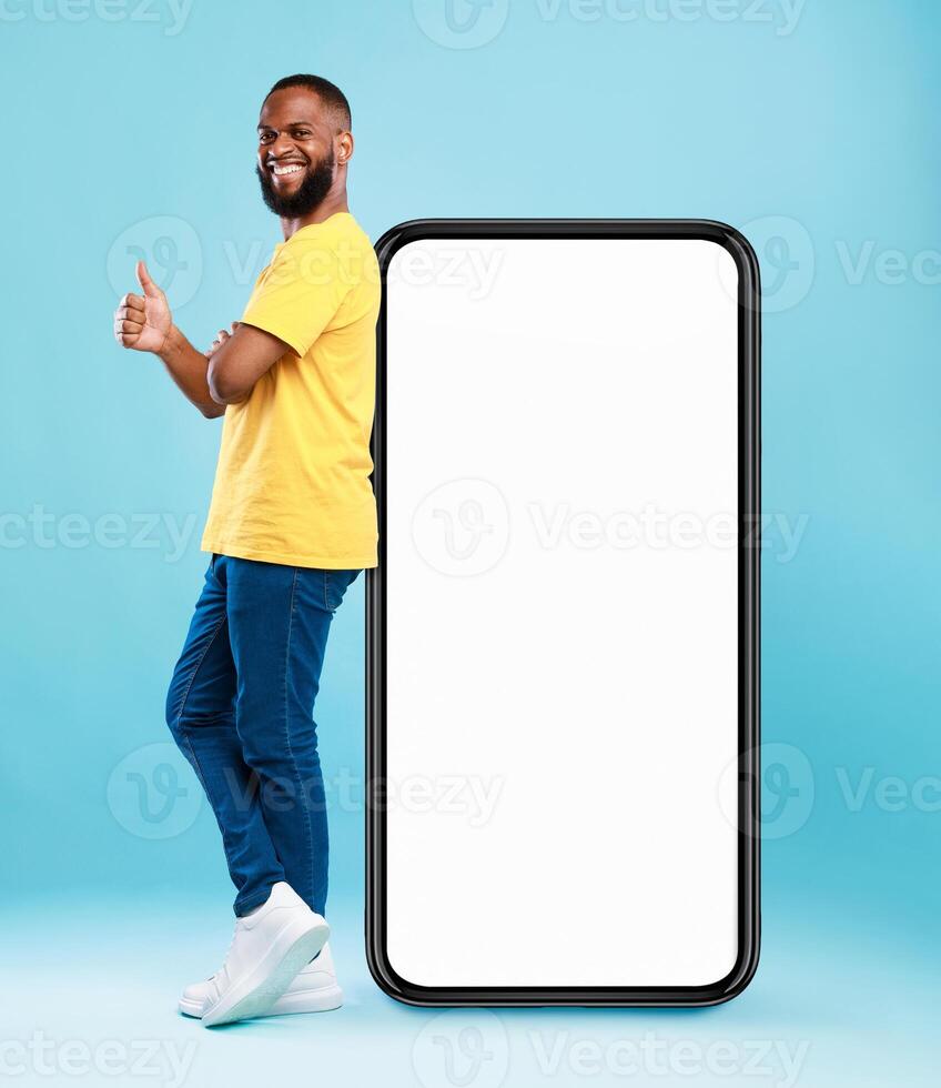 Funky black guy leaning on giant cellphone, showing thumb up gesture, recommending new mobile app or cool website, mockup for your ad design. Blue studio background photo