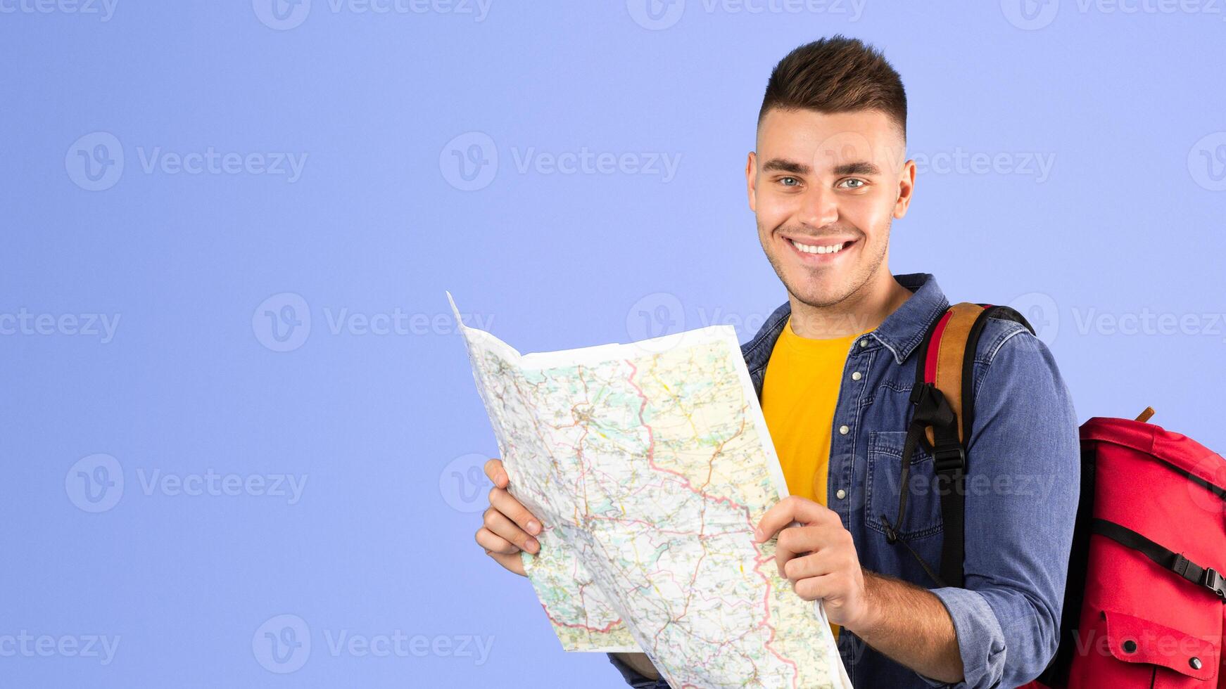 Positive young hiker with backpack and map planning his weekend camping trip route on blue studio background. Portrait of happy millennial traveler ready to go trekking photo