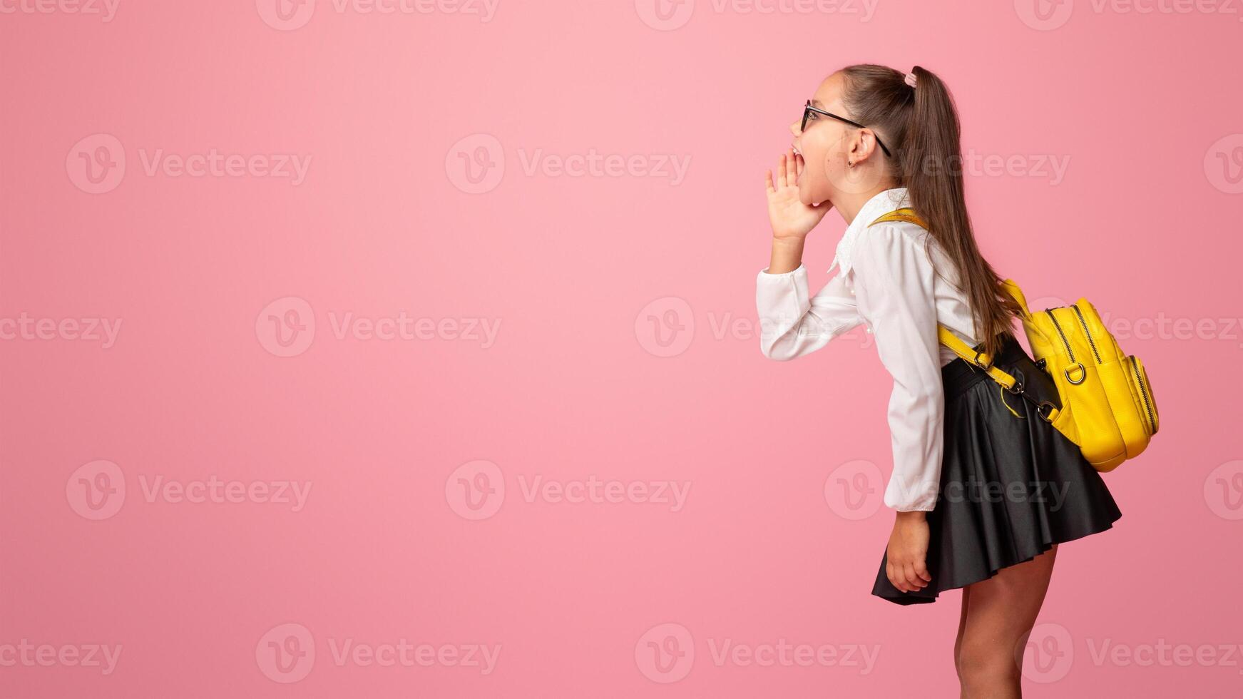 Shares a secret. Preschooler girl in uniform with backpack says whisper to abstract cloud for words on pink background, side view, studio shoot photo